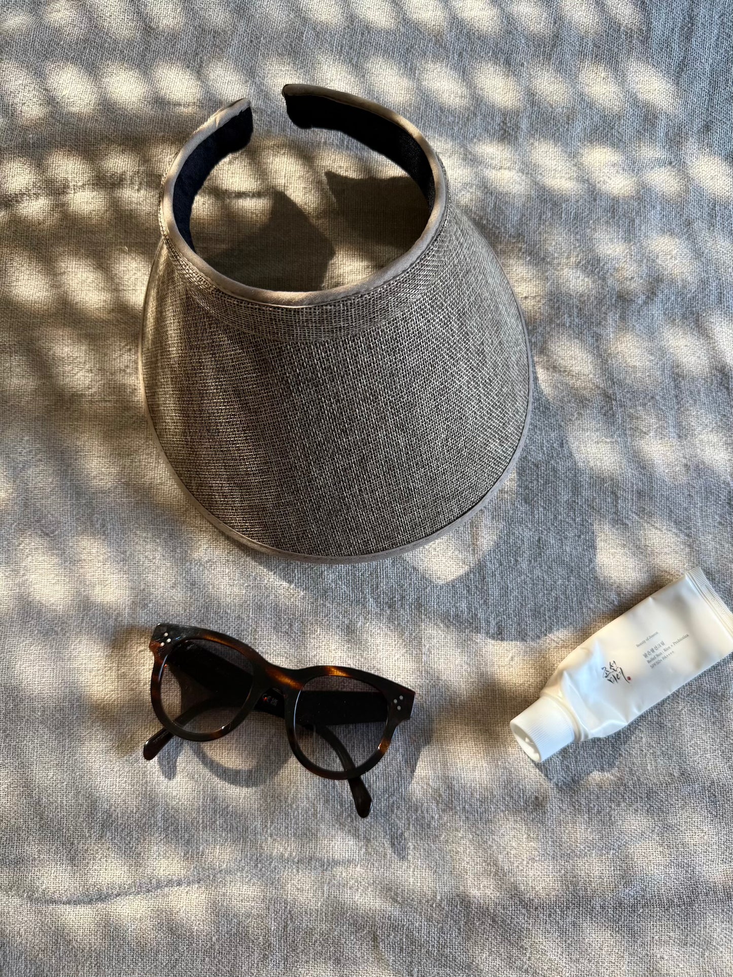 Gray visor, sunglasses, and sunscreen on a textured surface with sunlight casting shadows.